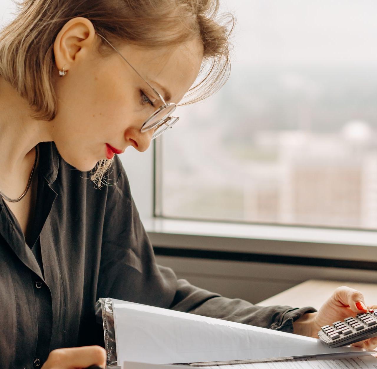 Businesswoman working with calculator and financial documents by window, focused and analytical.