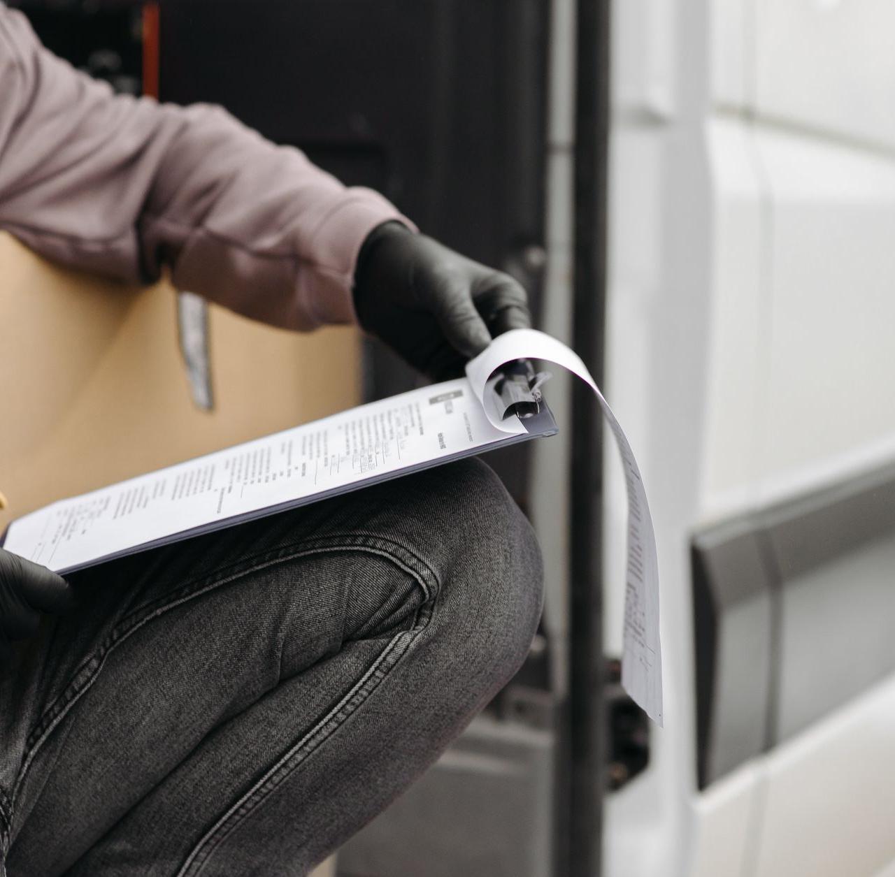 A delivery worker in gloves checking documents on a clipboard by a van.
