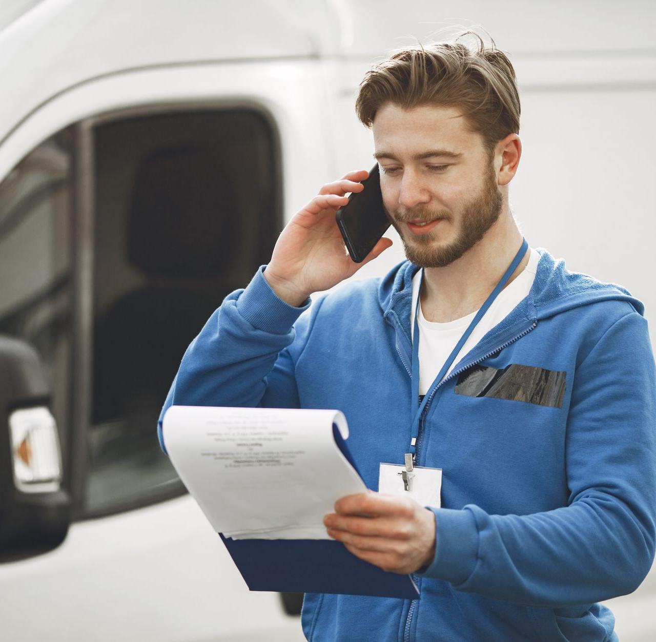 A delivery man in a blue hoodie makes a phone call while holding a clipboard, standing by a white van.