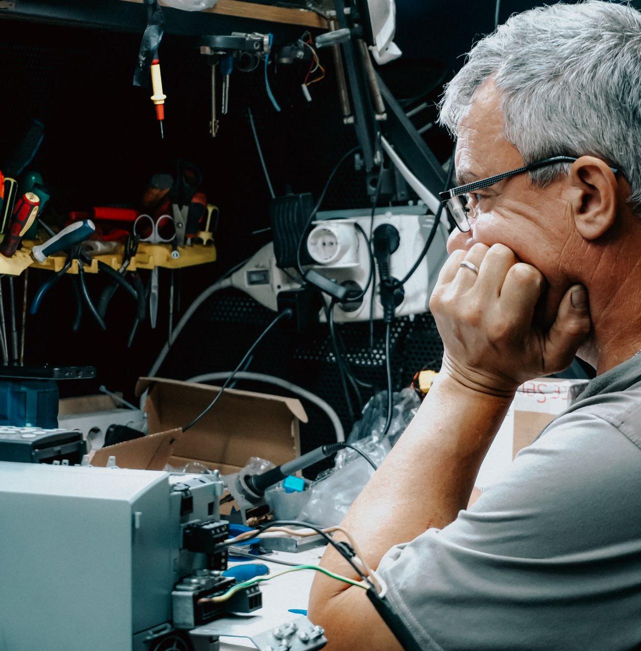 An adult man inspects electronic equipment at a cluttered workbench, deep in thought. Indoor electronics repair.