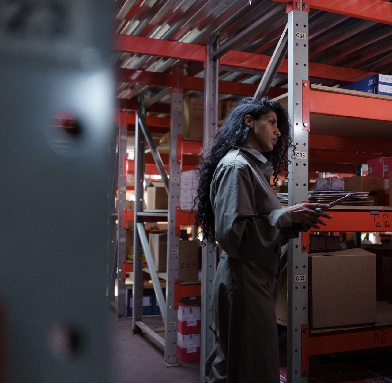 An adult woman in uniform checking inventory in a warehouse aisle.