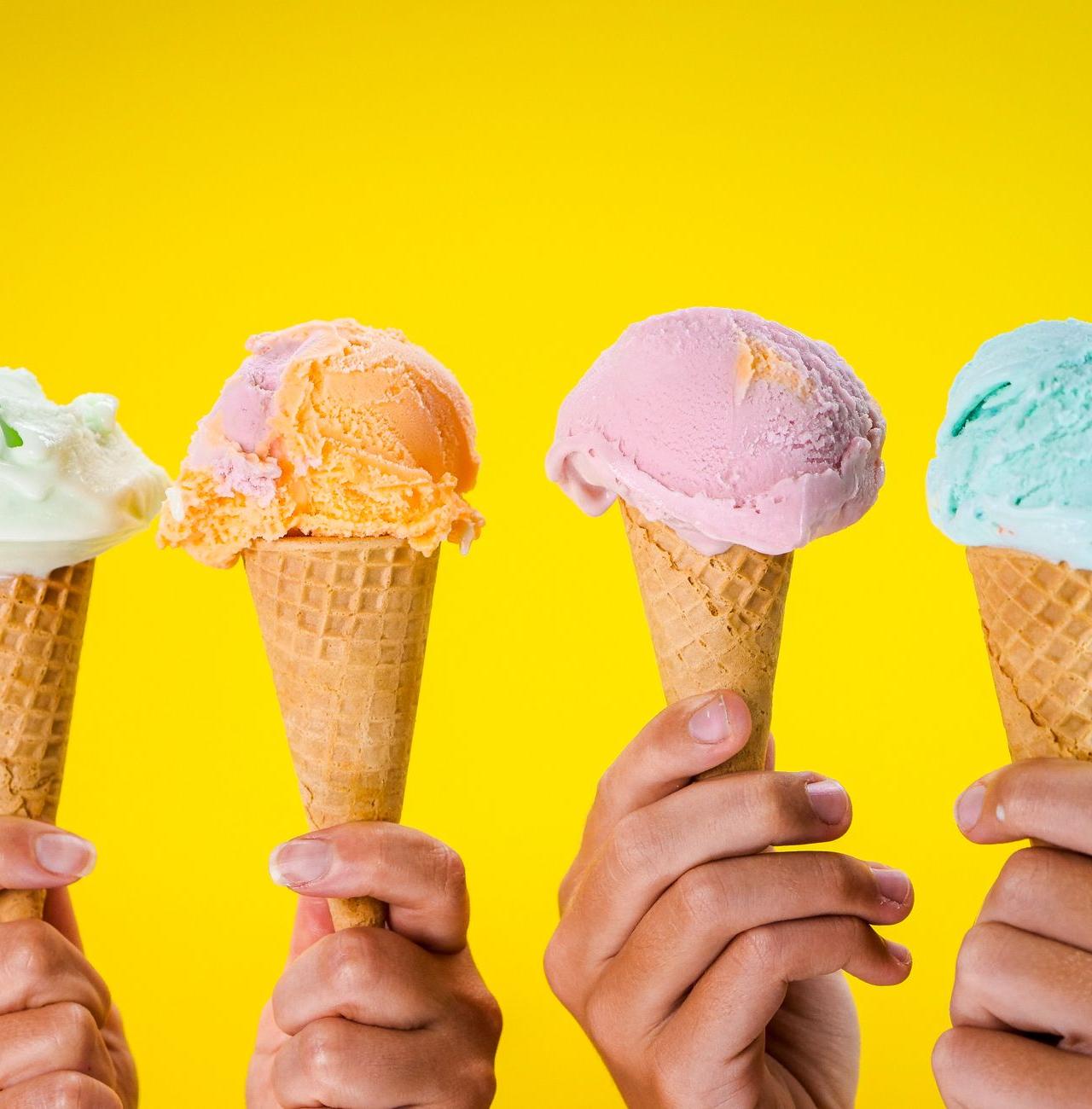 Four colorful ice cream cones held up against a bright yellow backdrop, showcasing various flavors.
