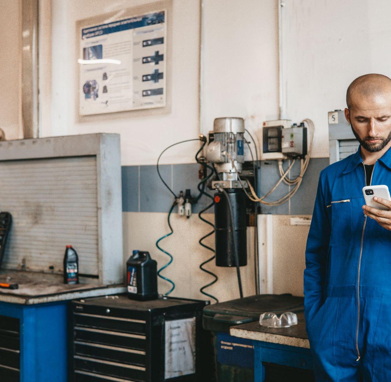Mechanic in blue coveralls using a smartphone in an auto repair workshop.