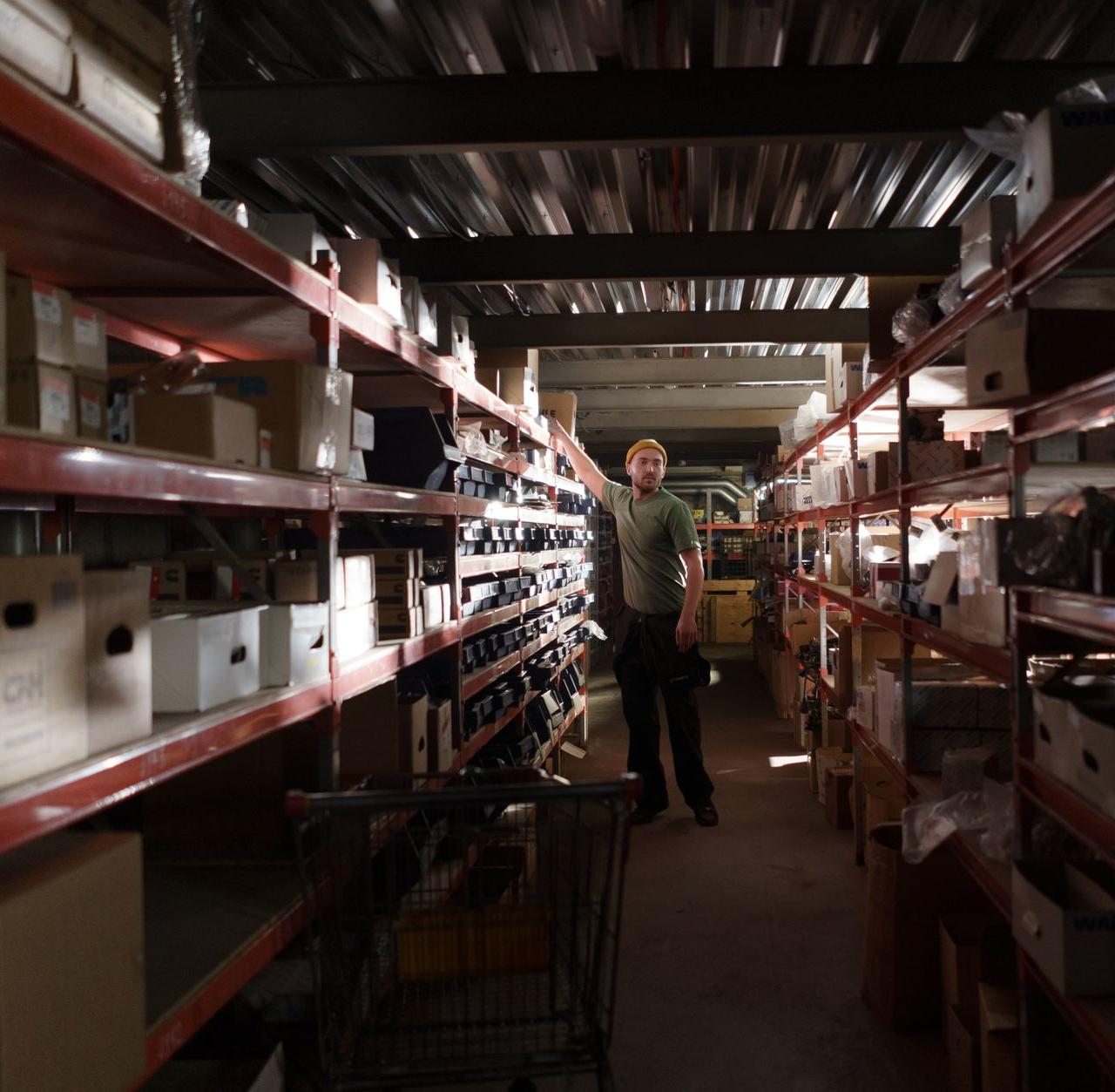A worker organizing inventory in a dimly lit warehouse aisle full of shelves and boxes.