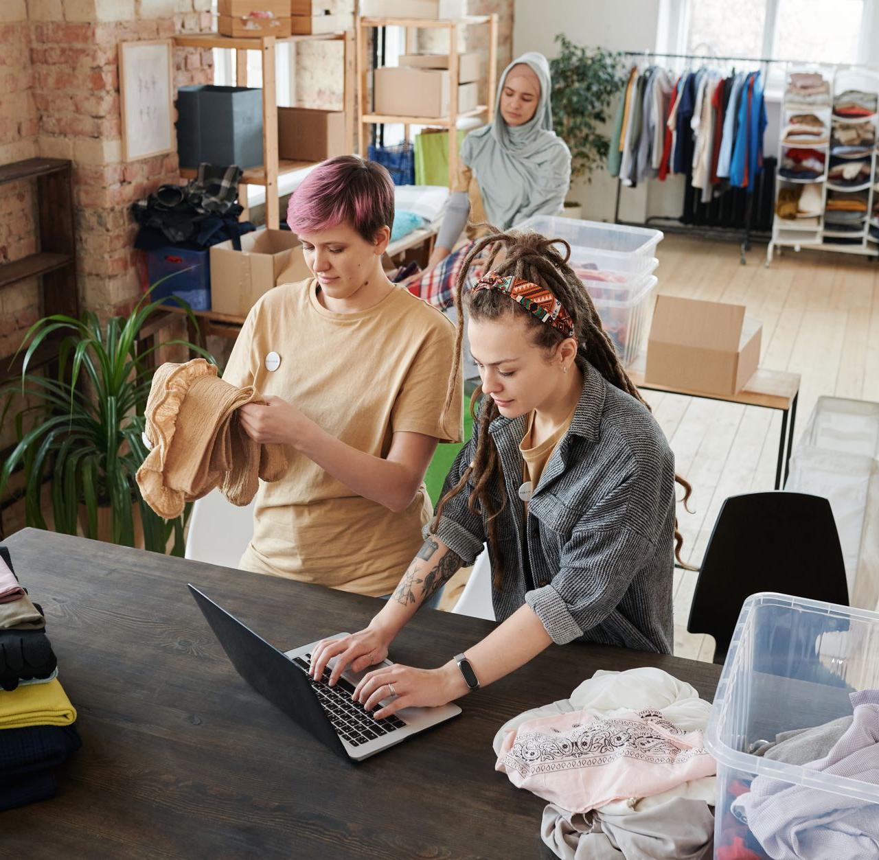 Women organizing clothes and using a laptop in a donation center. Diverse teamwork and collaboration.