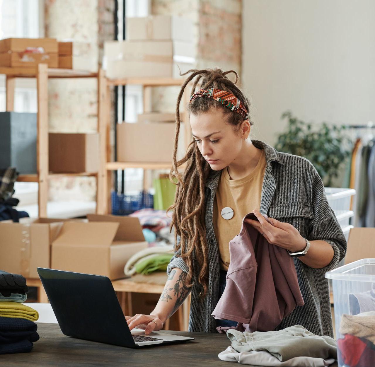 Woman with dreadlocks using a laptop while sorting clothes in a loft for volunteer work.