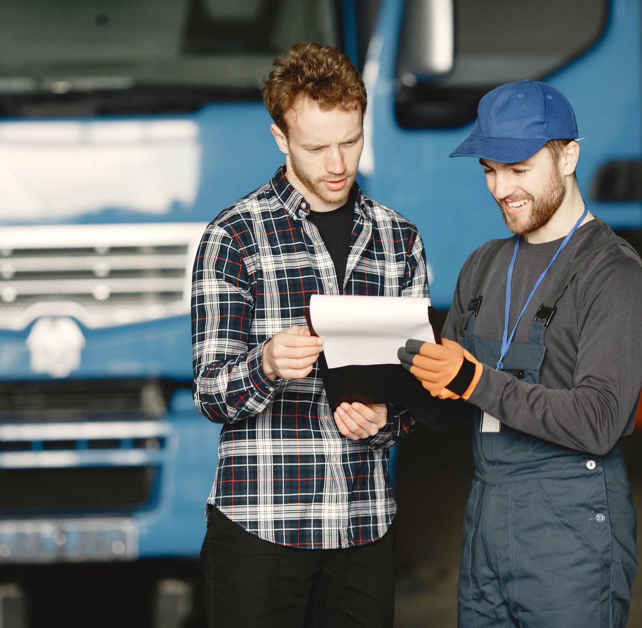 Two men in discussion over vehicle maintenance documents in a garage setting with trucks in the background.