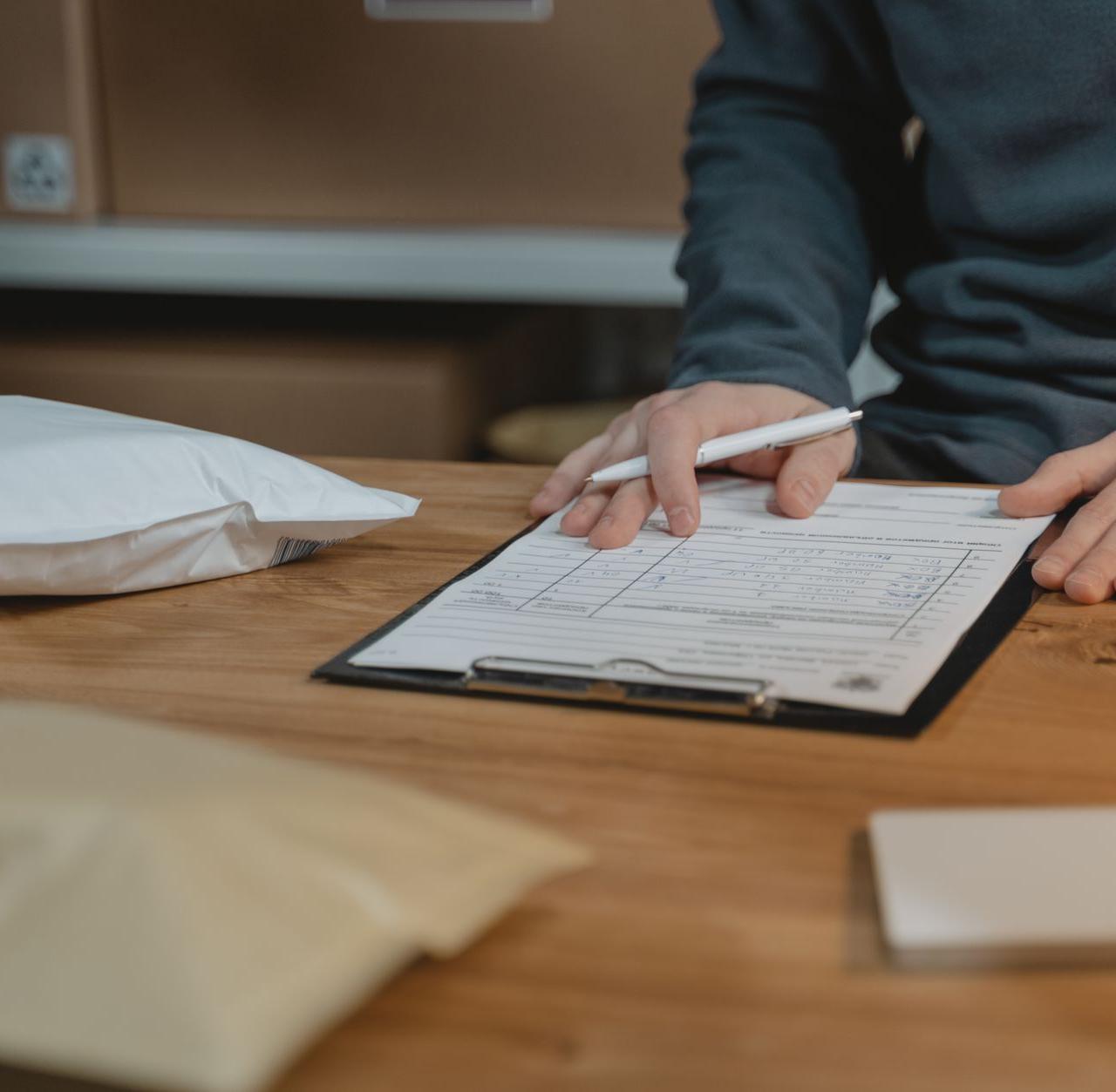 Close-up of a person filling out a form on a clipboard with packages nearby in an office setting.