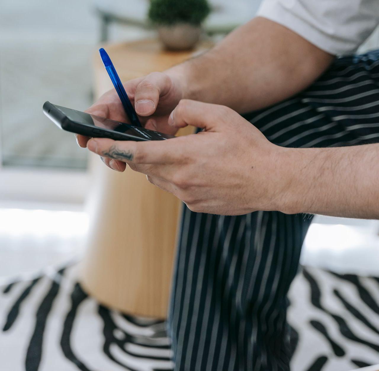 Man in striped pants texting on a smartphone with a blurry background inside a modern room.