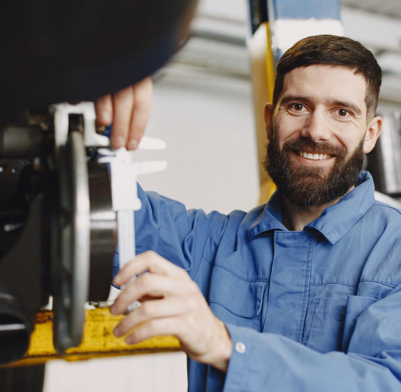 Mechanic working on a car in a garage, wearing blue overalls and smiling confidently.