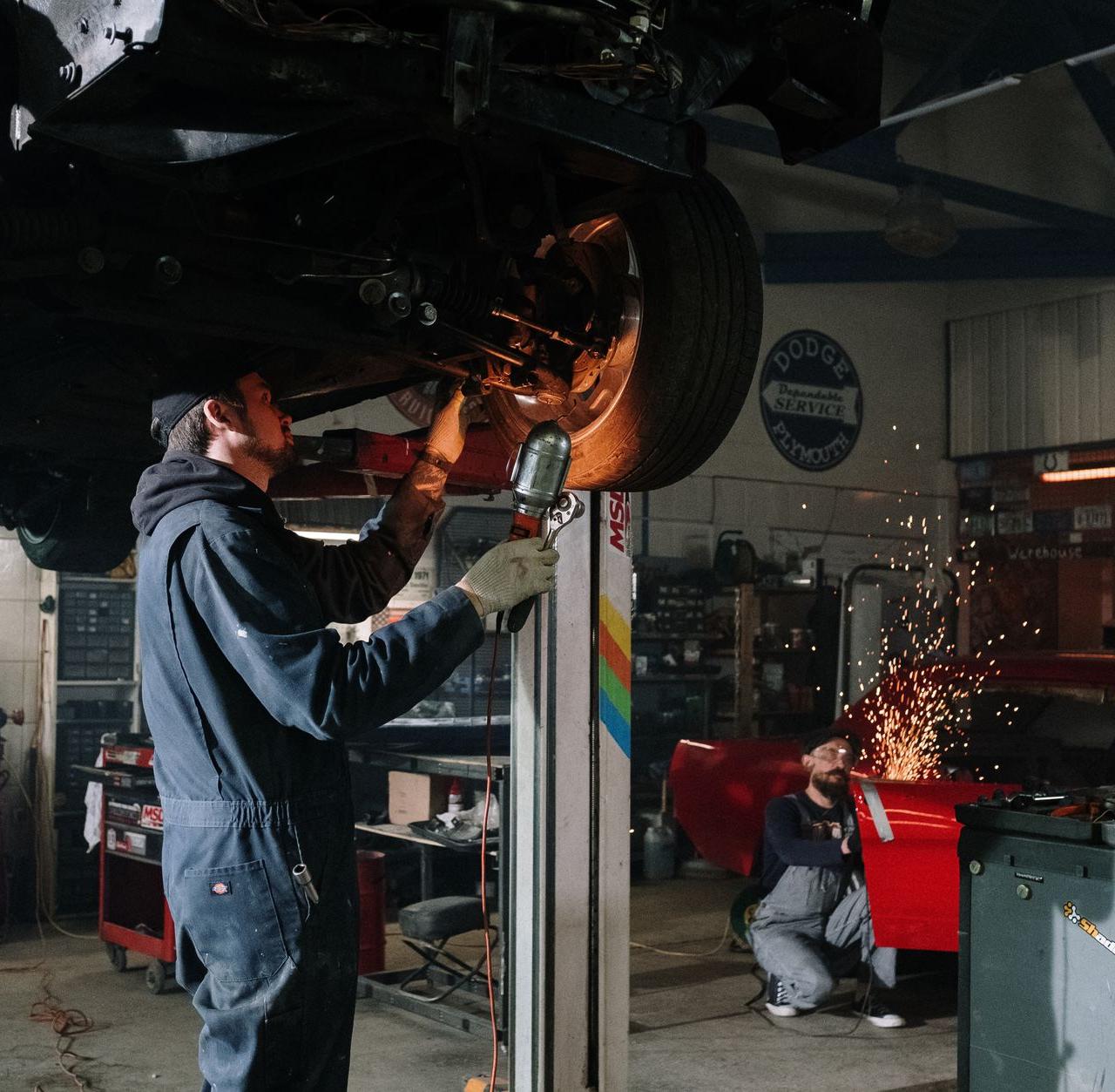 Two automotive technicians repairing a car in a busy workshop with sparks flying.