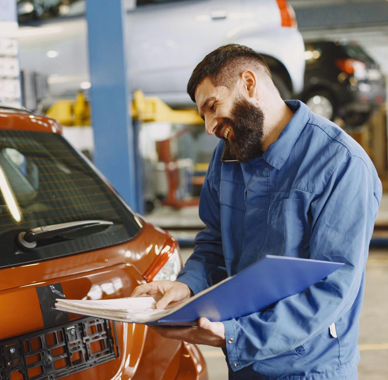 Smiling auto mechanic inspects a car in a service garage, wearing coveralls and holding documents.