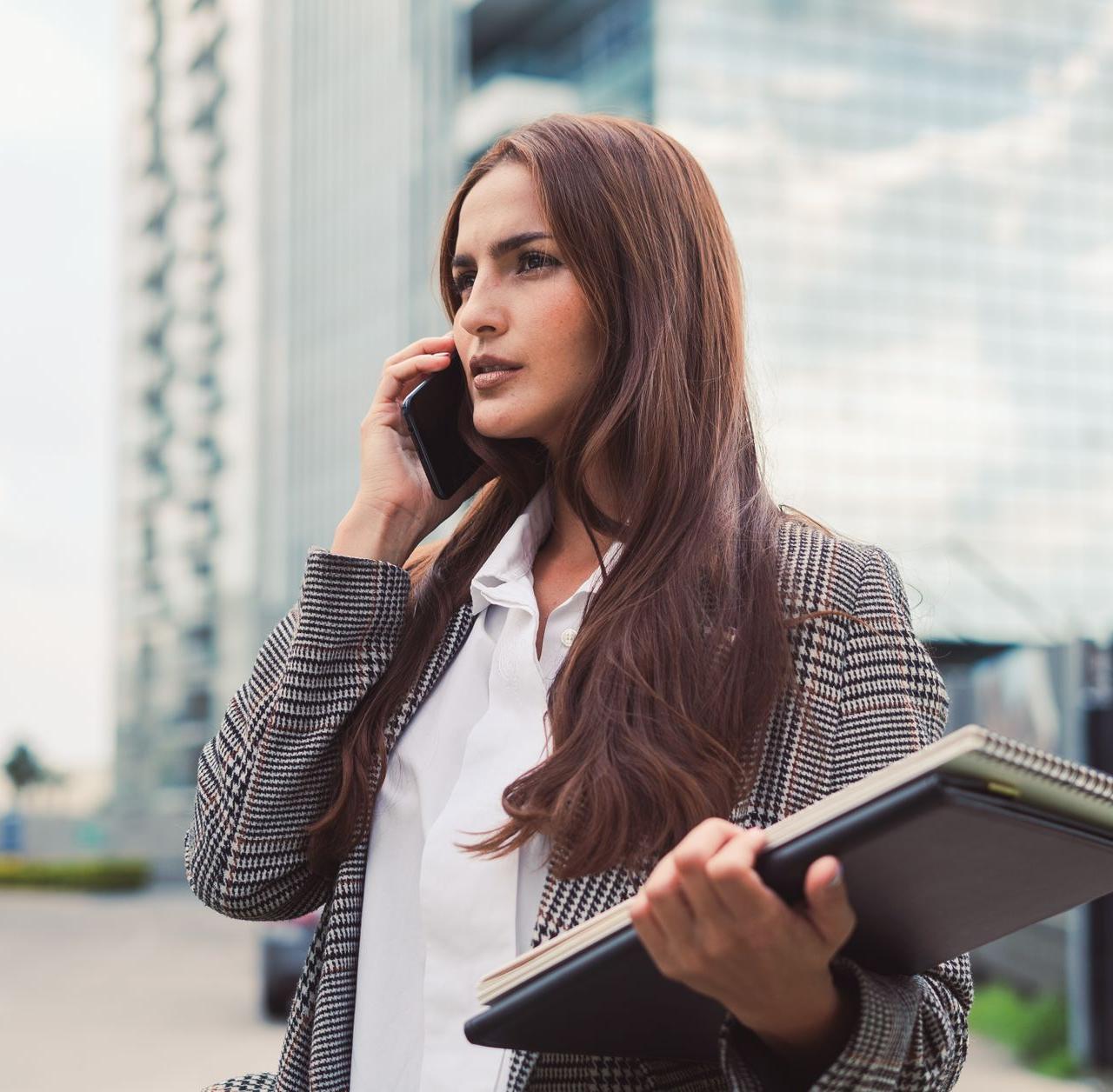 Confident businesswoman with long brown hair talking on phone outdoors, holding a notebook.