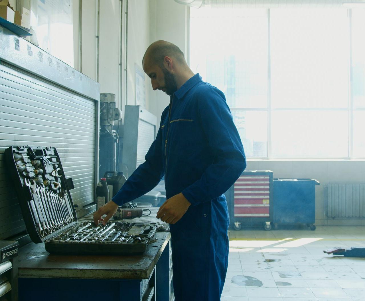 Mechanic inspecting tools in a workshop, wearing a blue jumpsuit with bright natural light from windows.