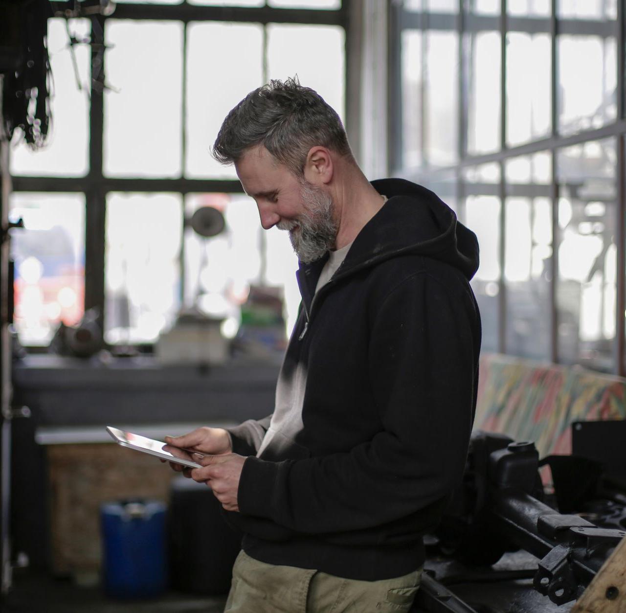Side view of positive adult bearded male mechanic wearing black clothes standing near workbench and using tablet while working in spacious workshop