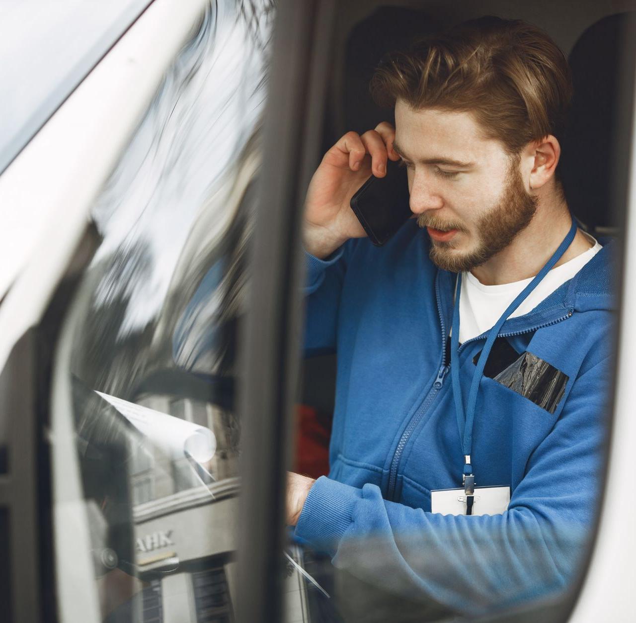 Bearded man in a blue hoodie uses smartphone inside vehicle, ID lanyard visible. Professional setting.