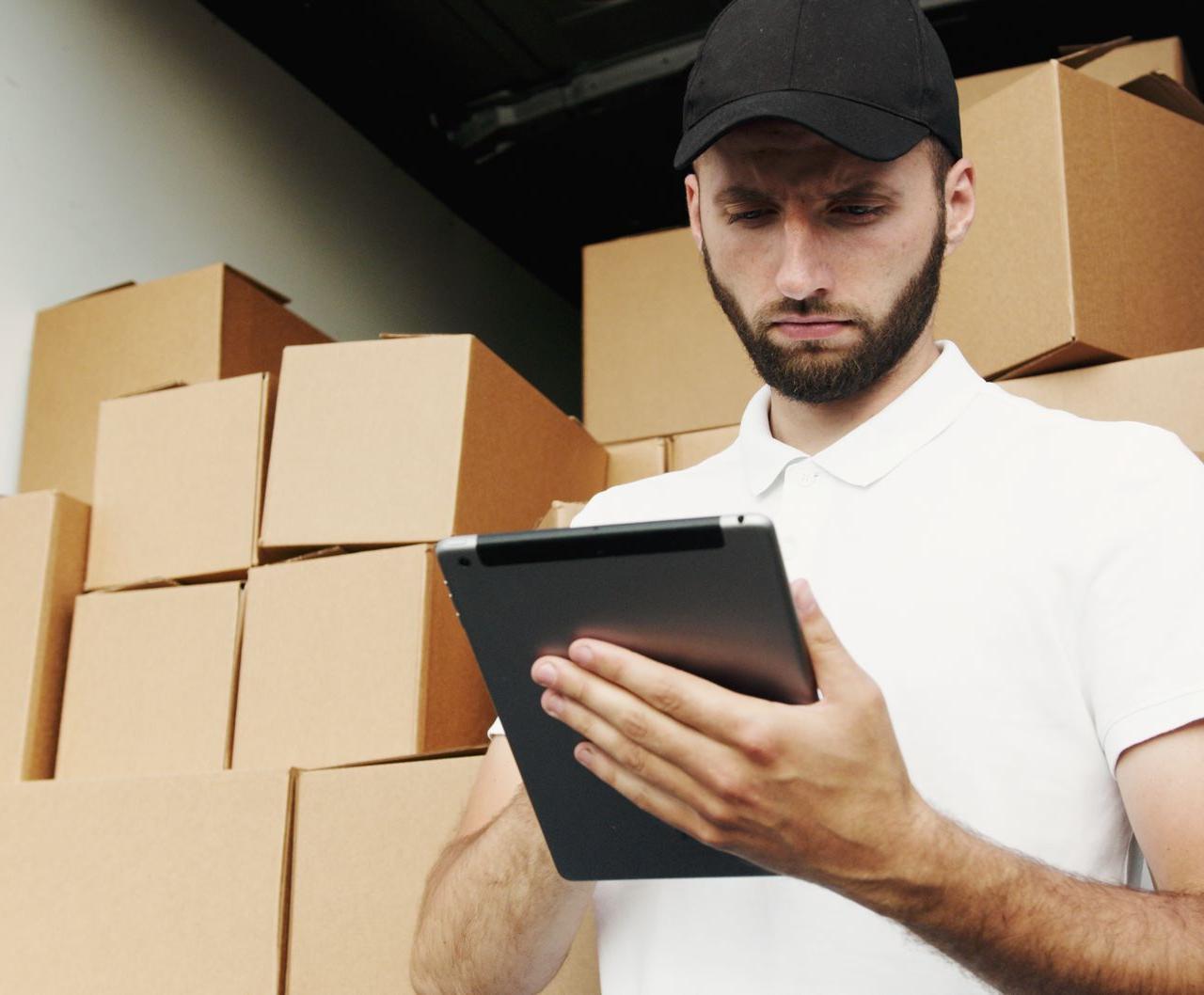 Delivery worker using a tablet to manage shipments with stacked boxes in the background.