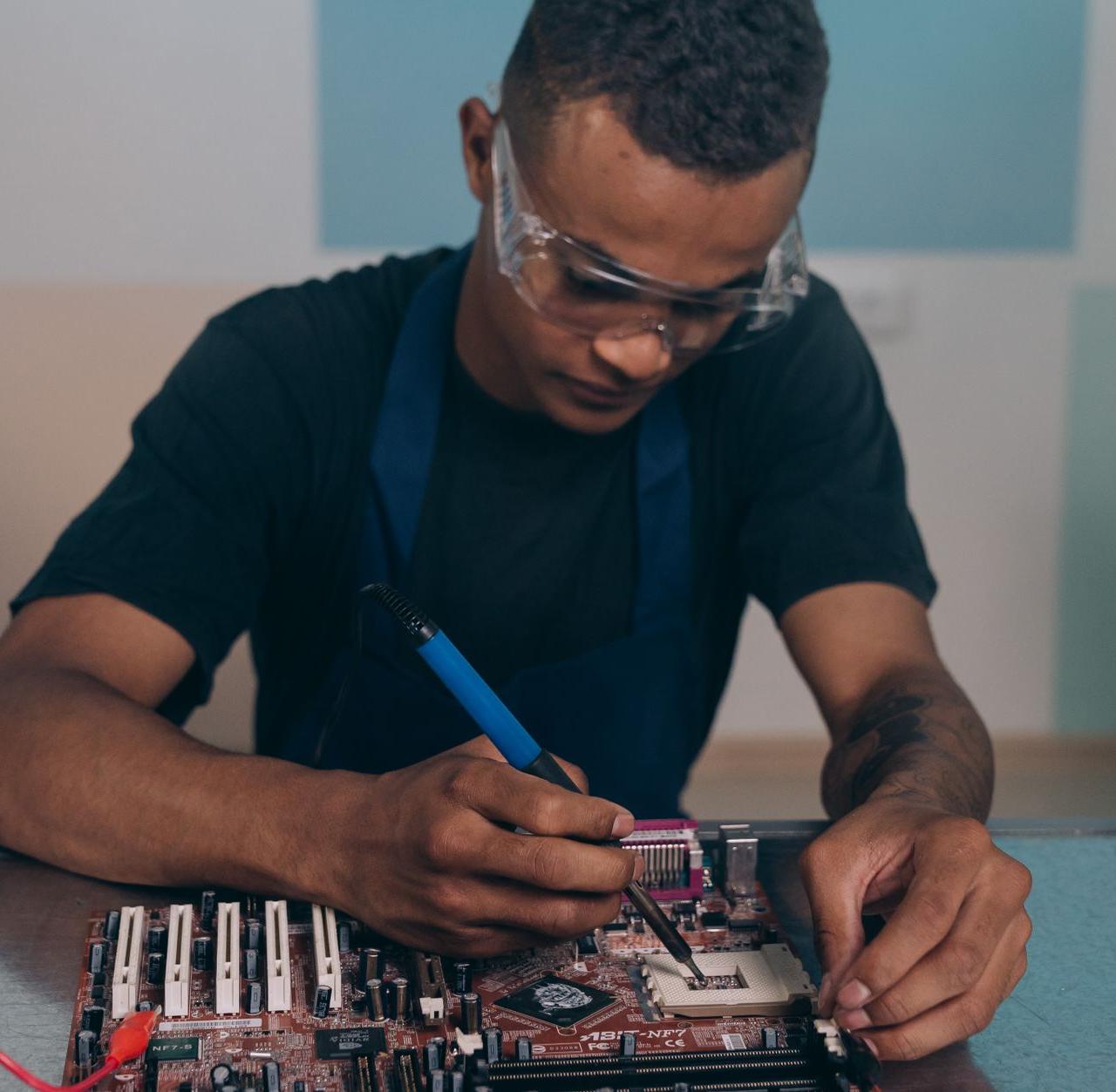 A technician wearing safety glasses works intently on a computer motherboard with a soldering iron.