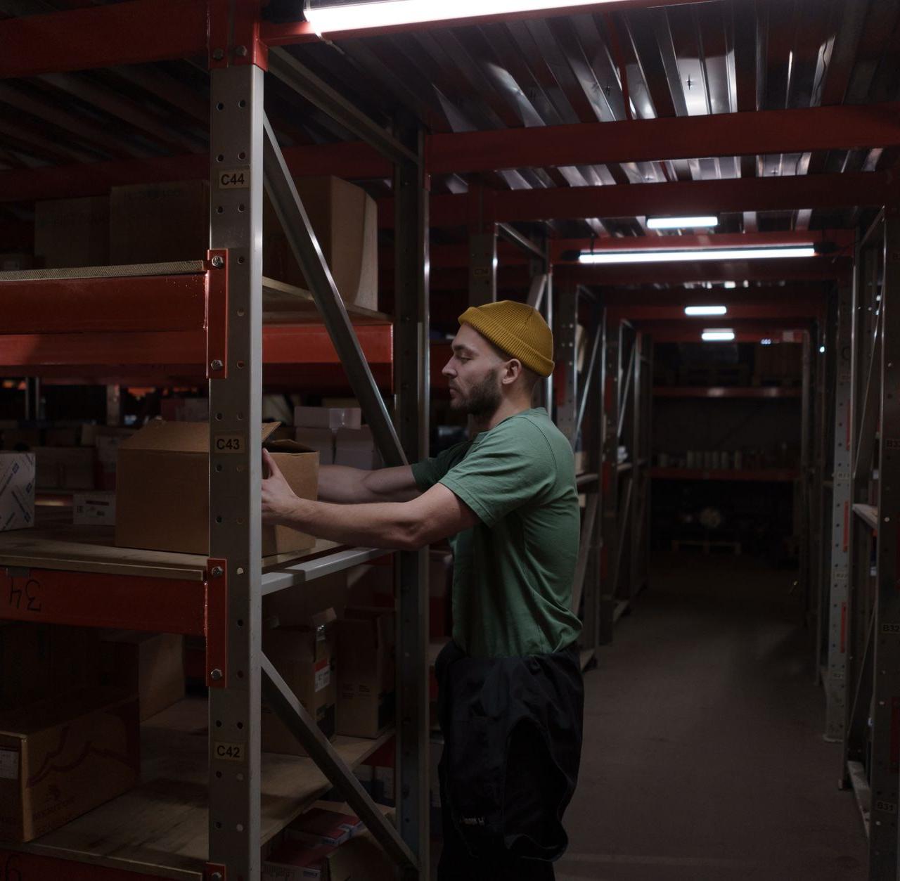 A man in a beanie and green shirt arranging boxes on shelves in a dimly lit warehouse storeroom.