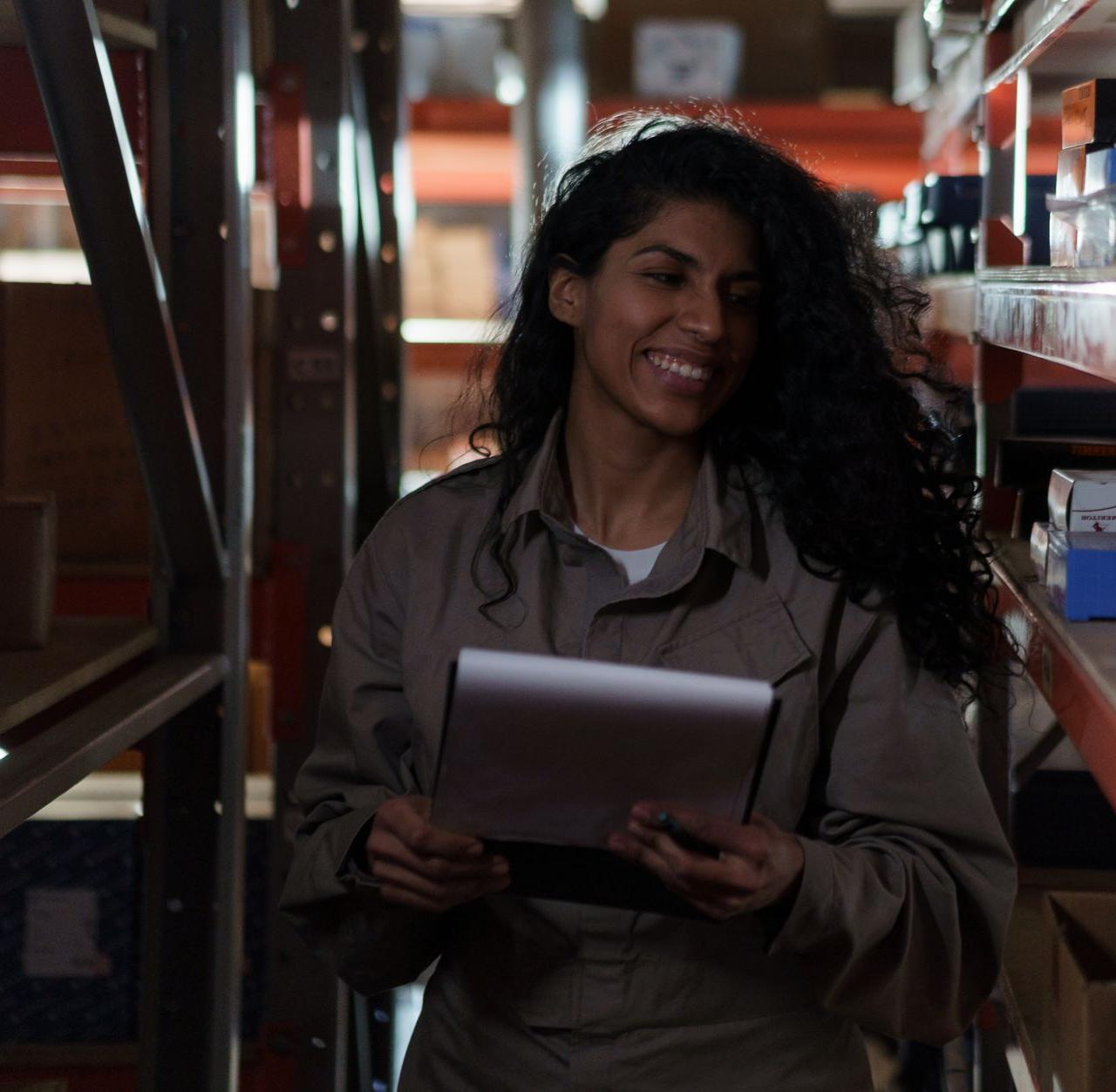 Woman managing inventory with clipboard, smiling in a dimly lit warehouse aisle.
