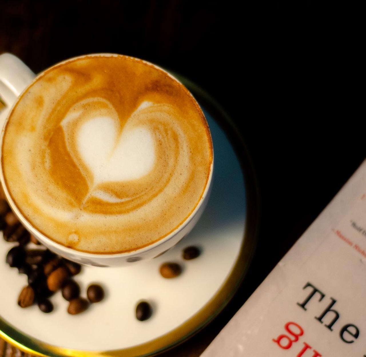 A cup of latte with heart art surrounded by coffee beans next to a book.
