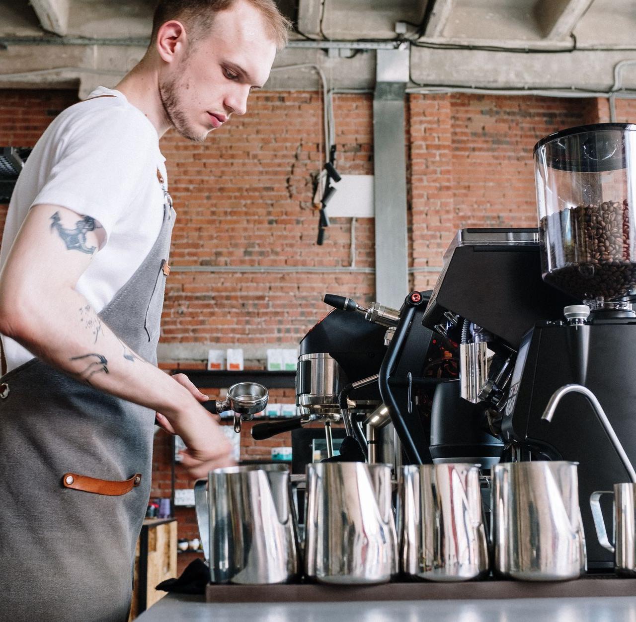 A young male barista preparing coffee in a modern industrial café setting with brick walls.