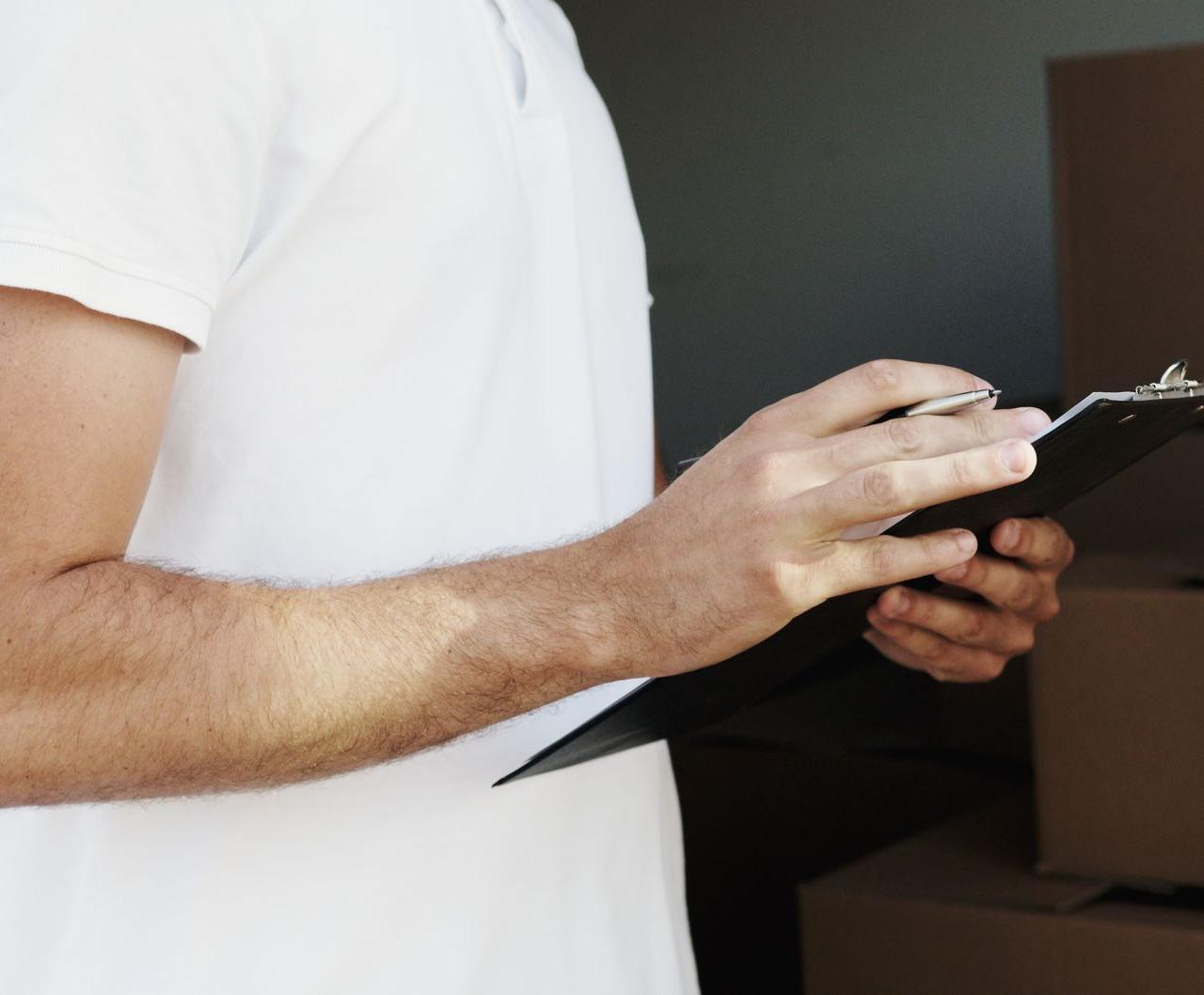 A delivery person in a white shirt holding a clipboard and pen near a vehicle with boxes.