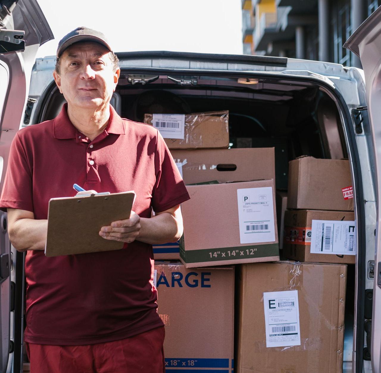 Elderly delivery person holds clipboard in front of open van filled with packages on a sunny day.