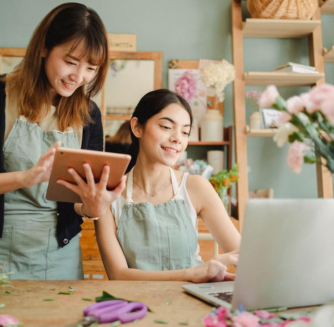 Cheerful multiethnic female coworkers wearing aprons at table with flowers browsing on tablet and netbook while working online in floristry studio
