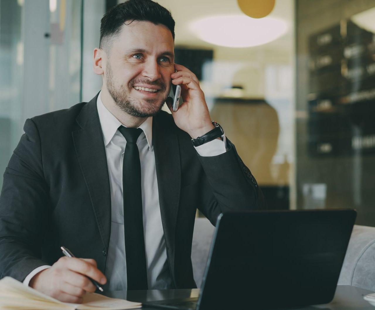 Professional man in a suit talking on the phone and working on a laptop indoors.