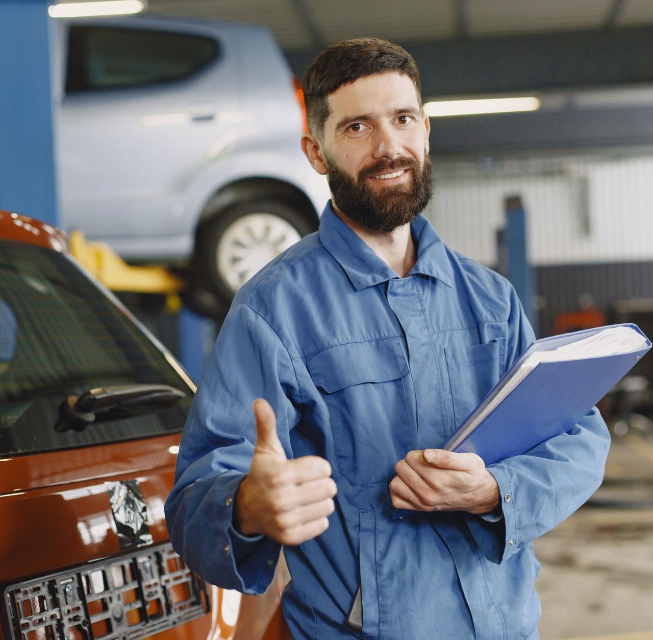 Auto mechanic in blue coveralls with clipboard giving thumbs up in garage.