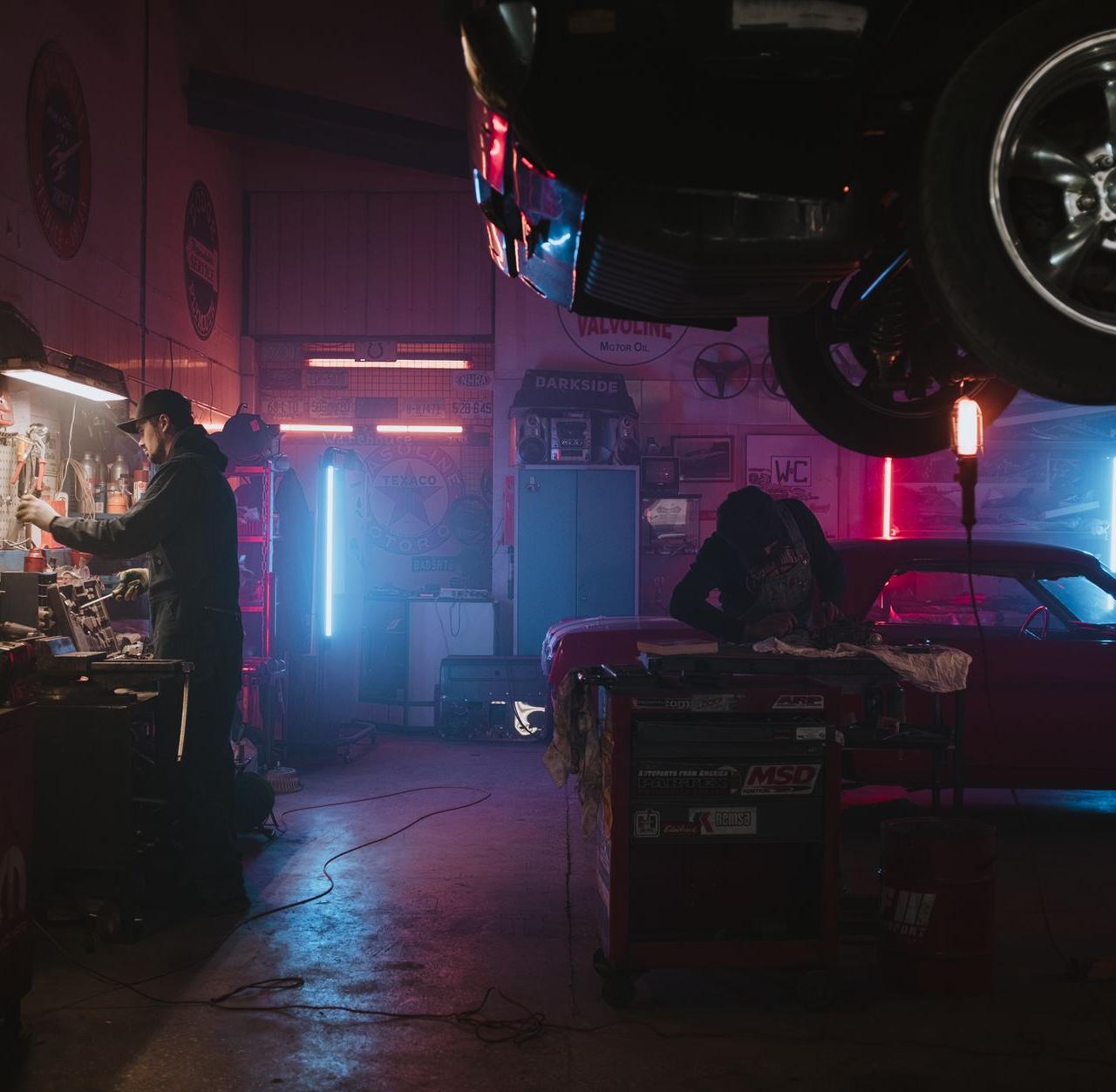 Mechanics working in a neon-lit garage, focusing on tools and car repairs under vibrant lights.