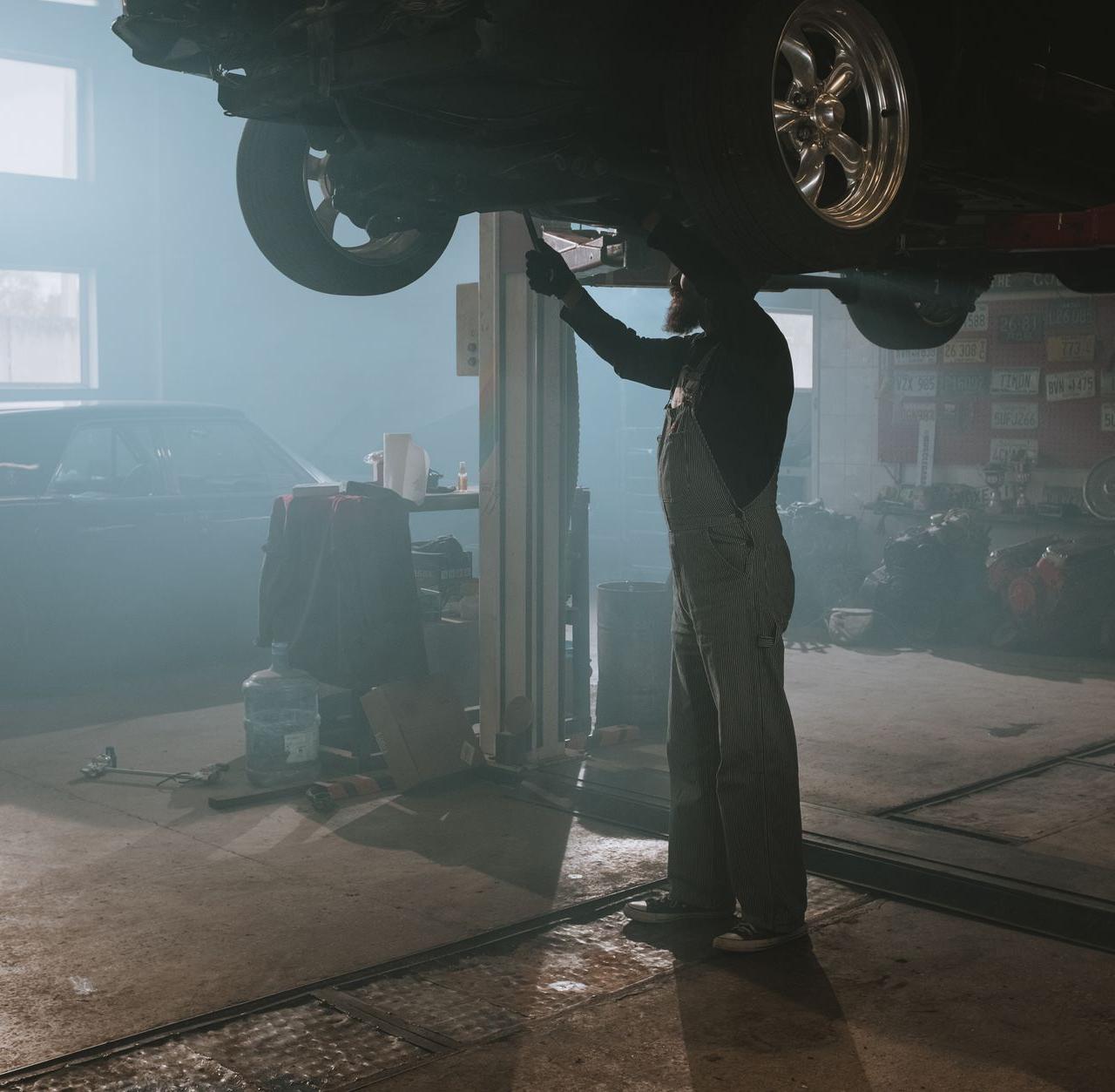 A mechanic is fixing a car raised on a lift in a dimly lit garage, creating a vintage ambiance.