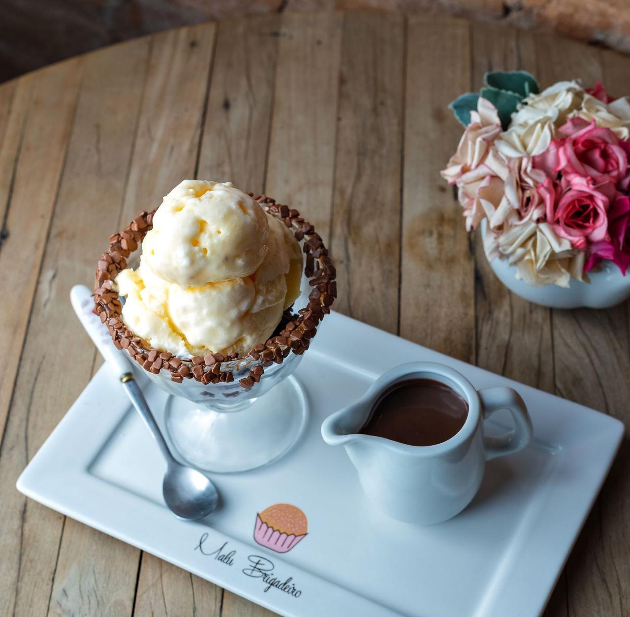 Scoops of ice cream served with chocolate sauce on a wooden table, alongside decorative flowers.
