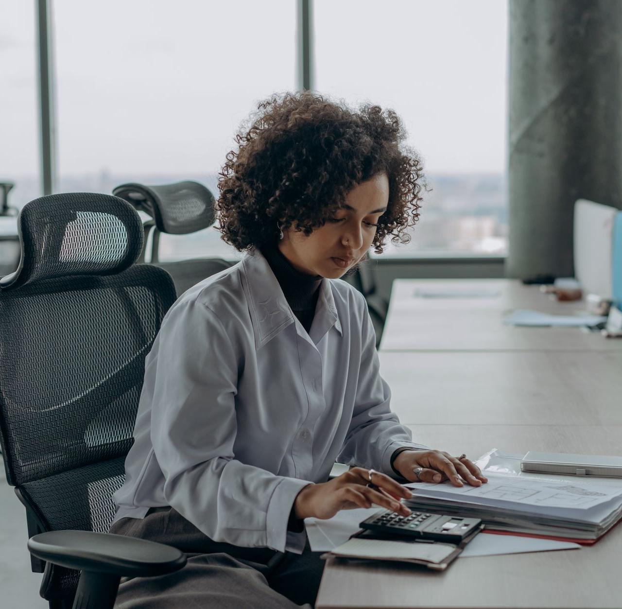 A professional woman with curly hair calculating documents in a modern office setting.