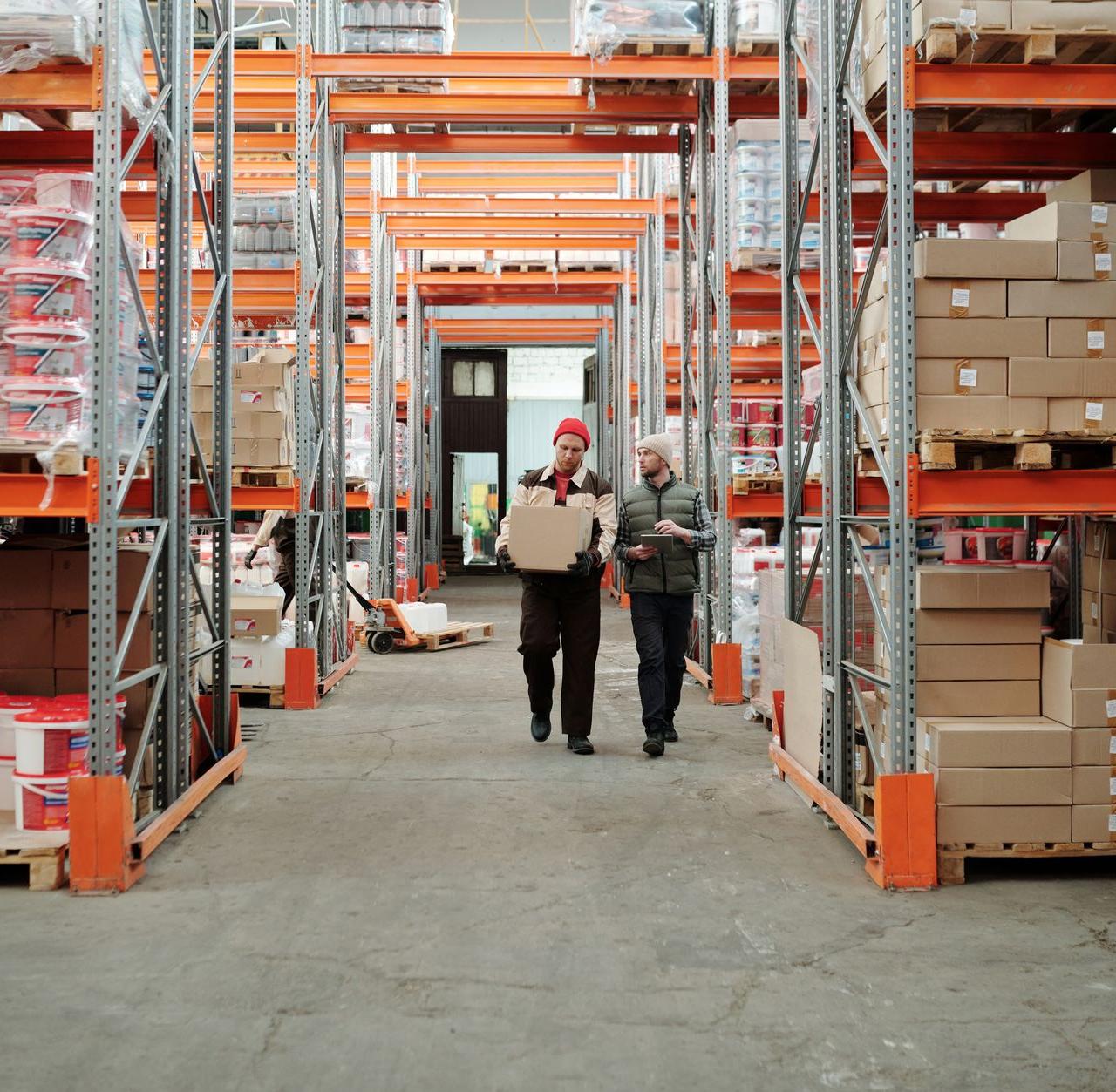 Two workers handle a package in a spacious warehouse surrounded by shelves stocked with boxes and products.
