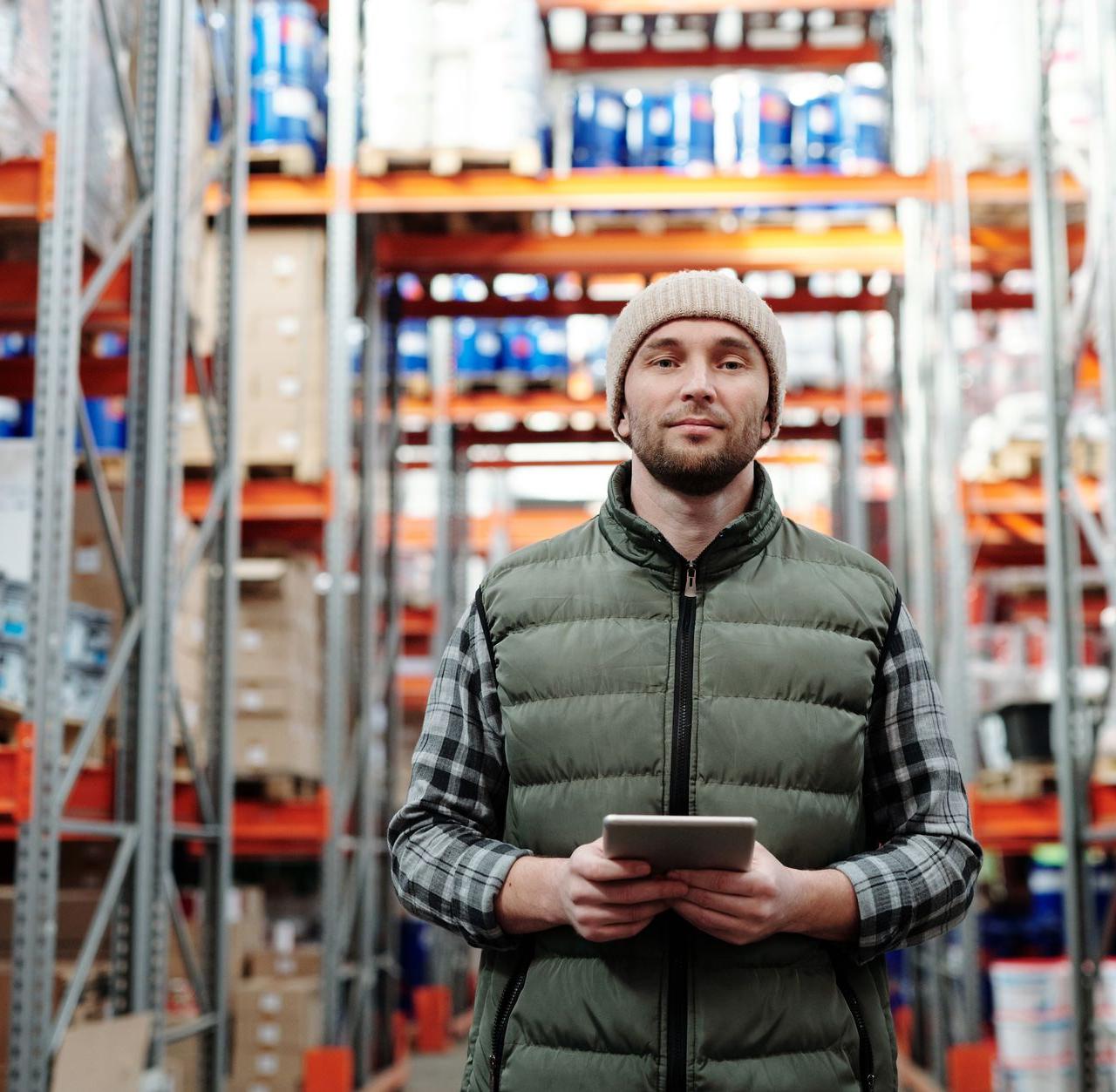 A warehouse worker wearing a beanie uses a tablet to manage inventory in a storeroom with shelves.