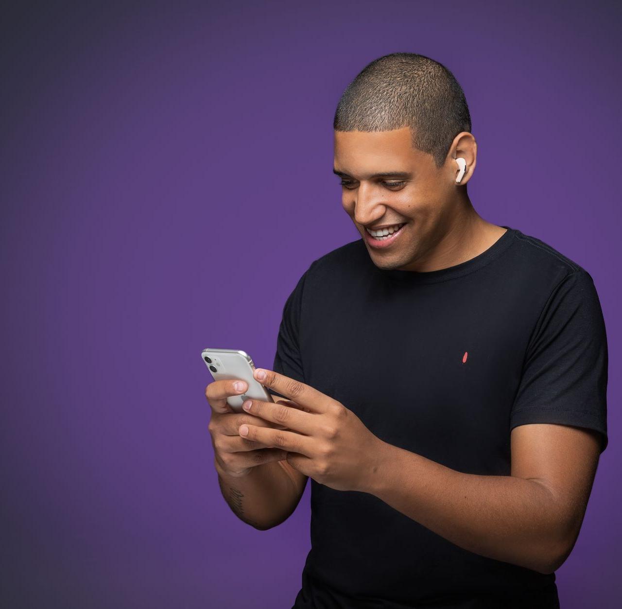 A cheerful man using a smartphone with earbuds on a violet backdrop.