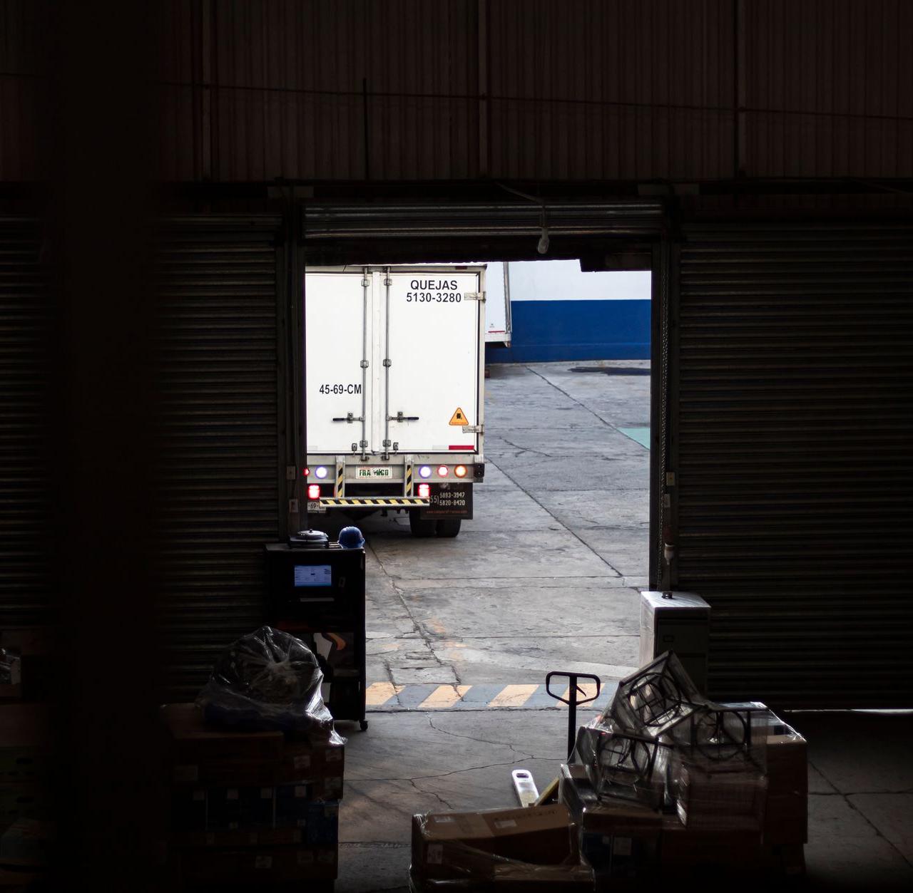 A white delivery truck exits a dimly lit warehouse loading dock, surrounded by packages and pallets.