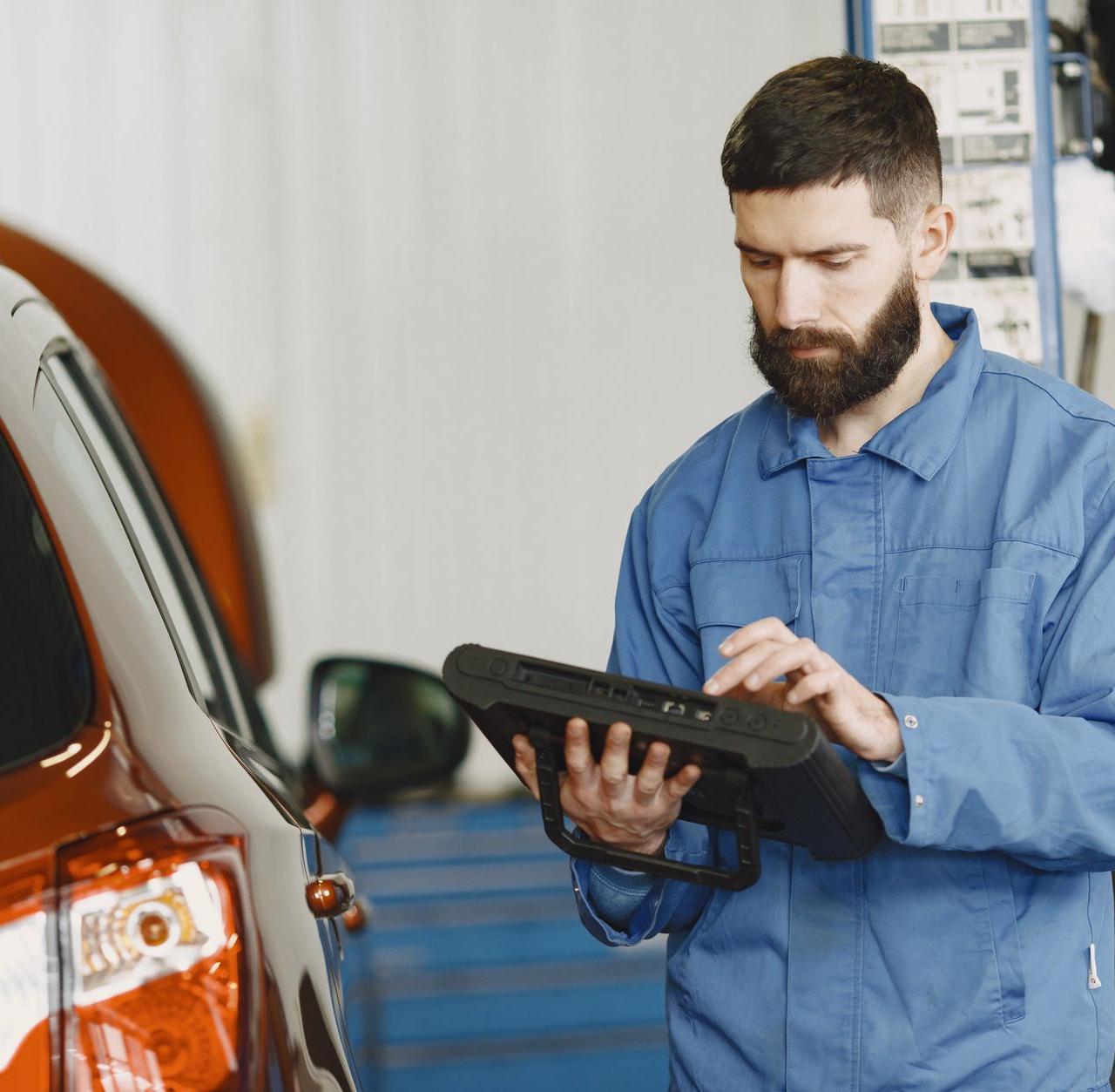 A mechanic in coveralls using a diagnostic tool at an auto repair shop beside a car.