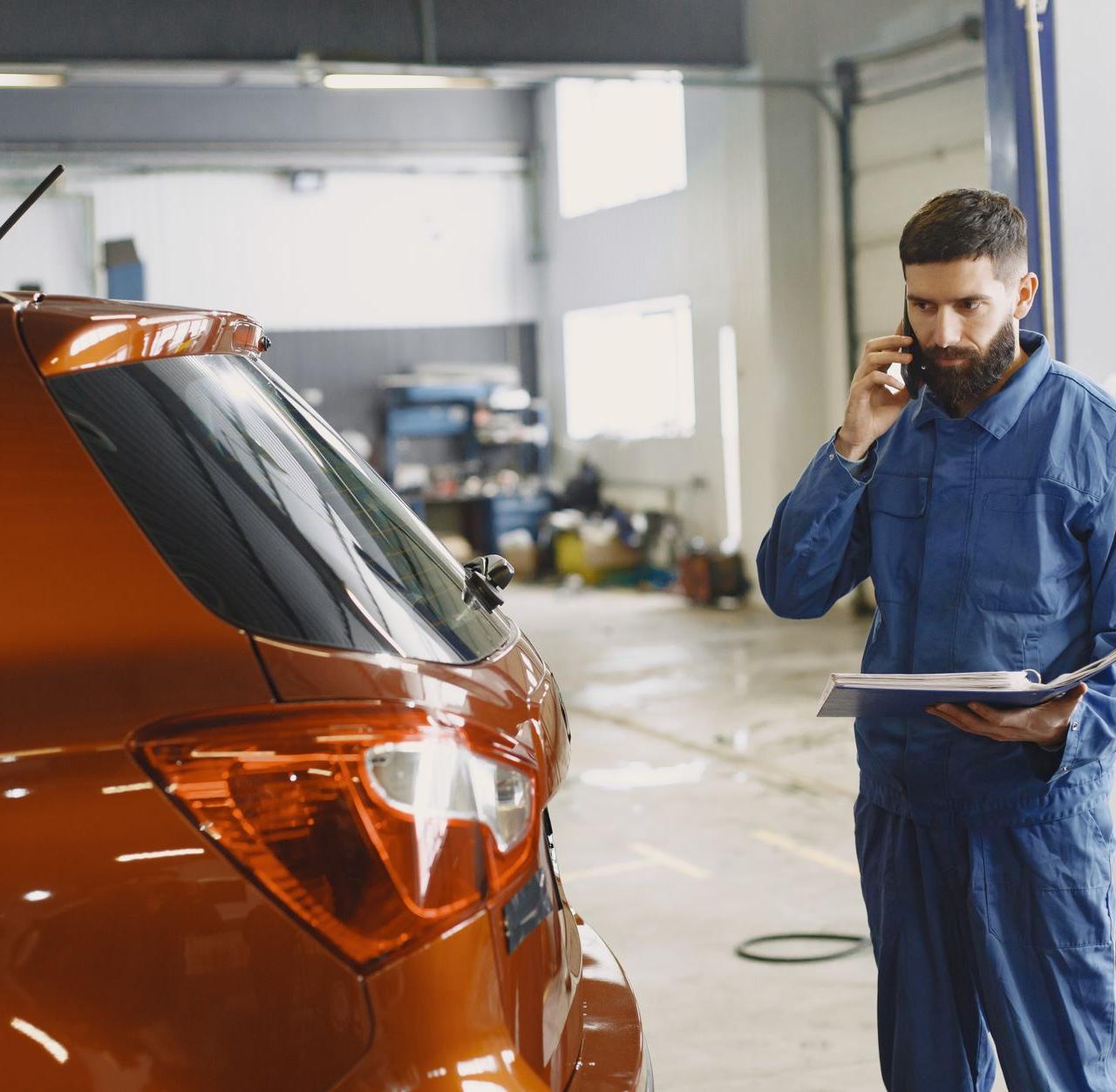 Mechanic in a blue uniform talks on phone beside a red car in an auto repair shop.