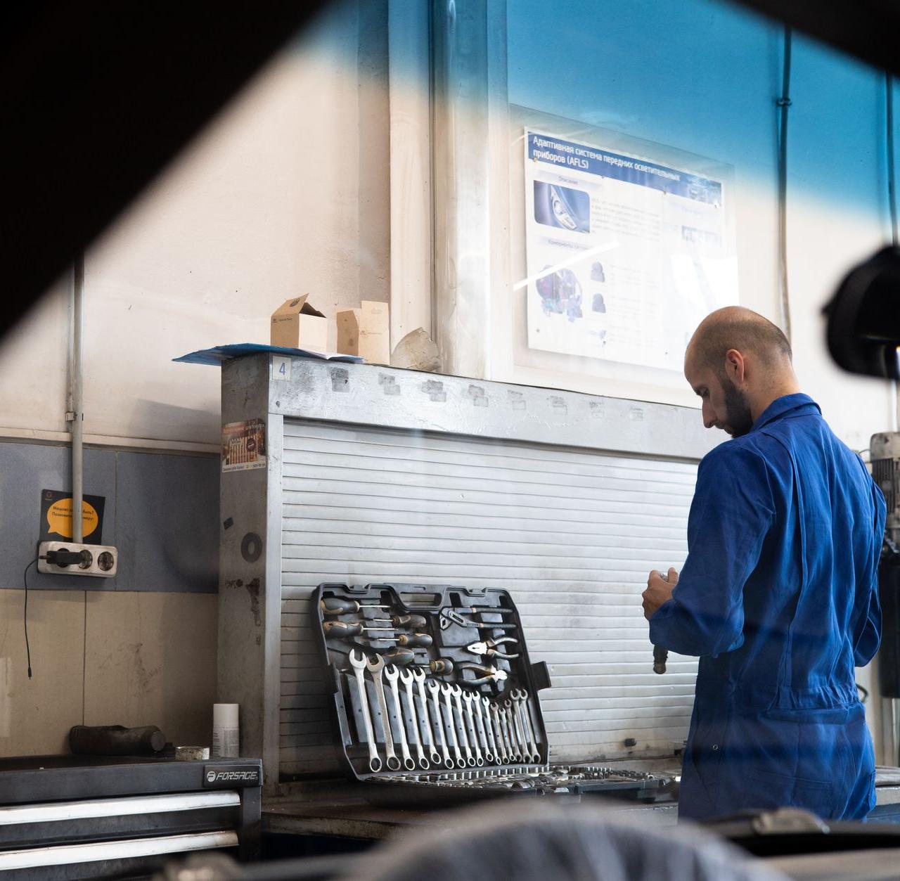 Bald mechanic in blue coveralls checking tools inside an automotive workshop.