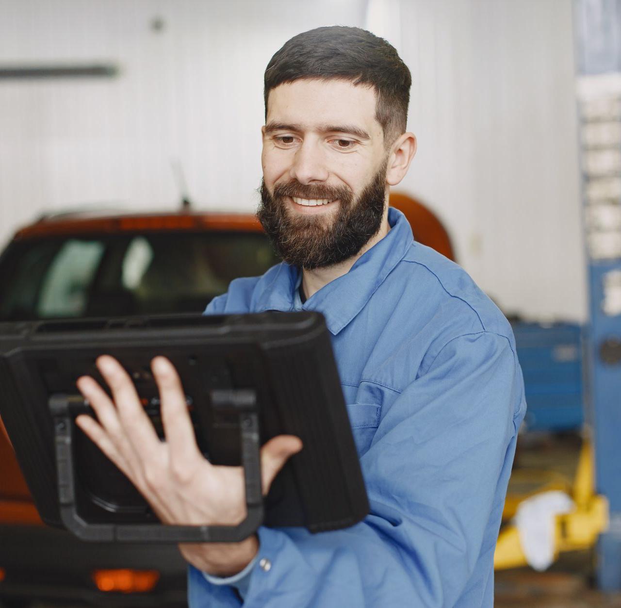 Smiling mechanic in blue uniform uses a digital tablet for vehicle diagnostics in a garage.