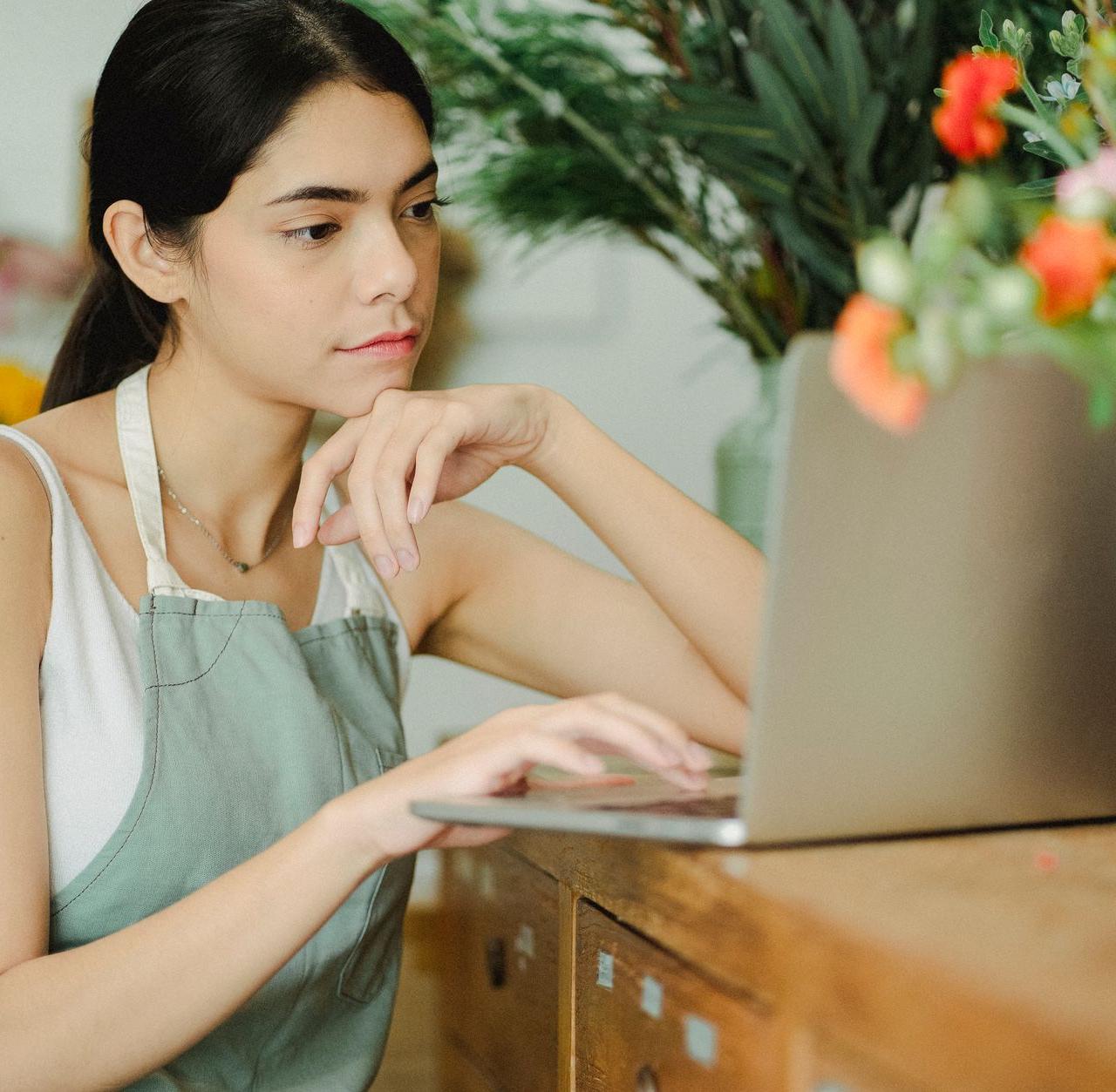 Concentrated female in apron sitting at wooden table with netbook and browsing while working in flower shop