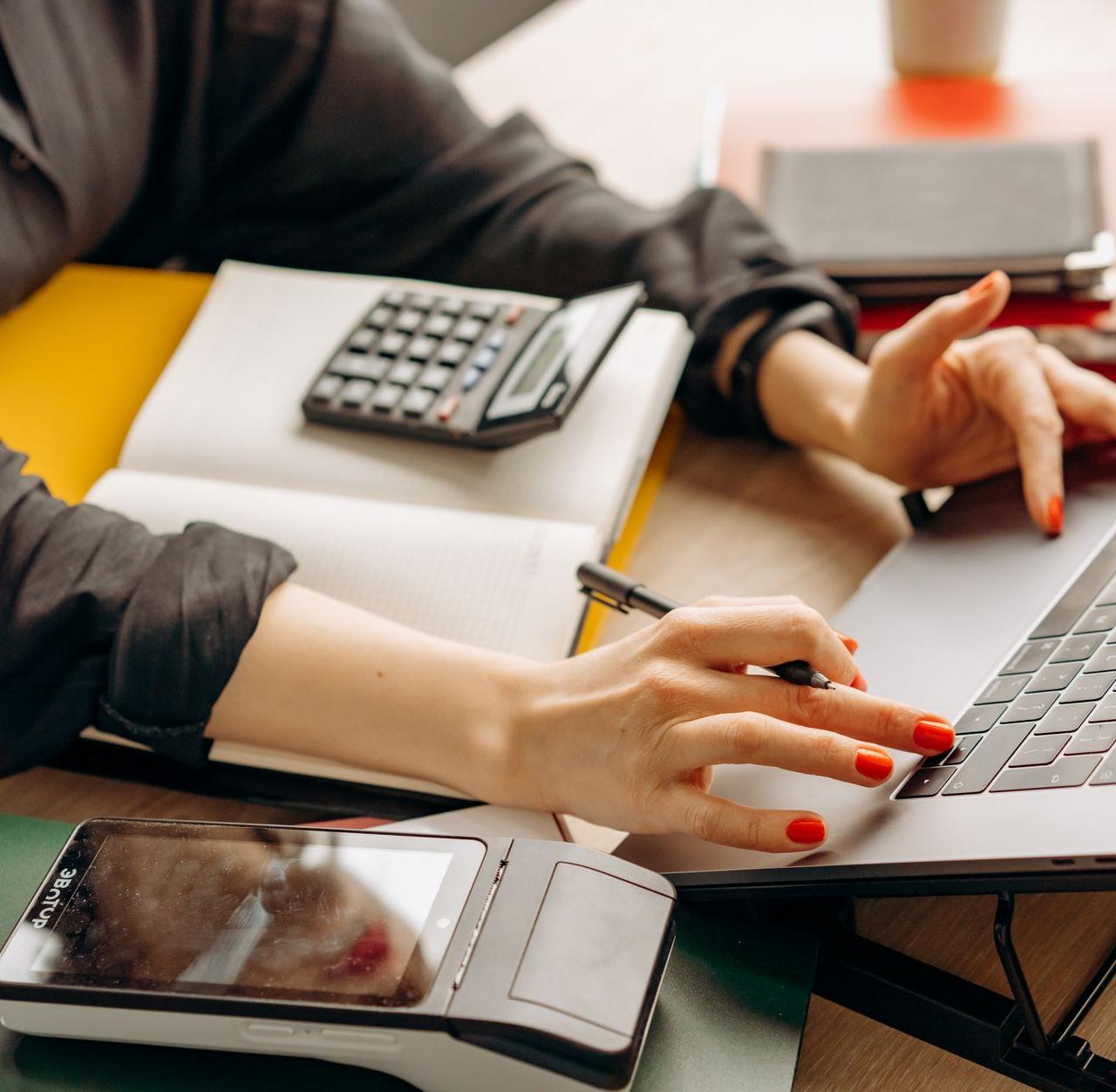 Close-up of a person using a laptop with a payment terminal and calculator on the desk.