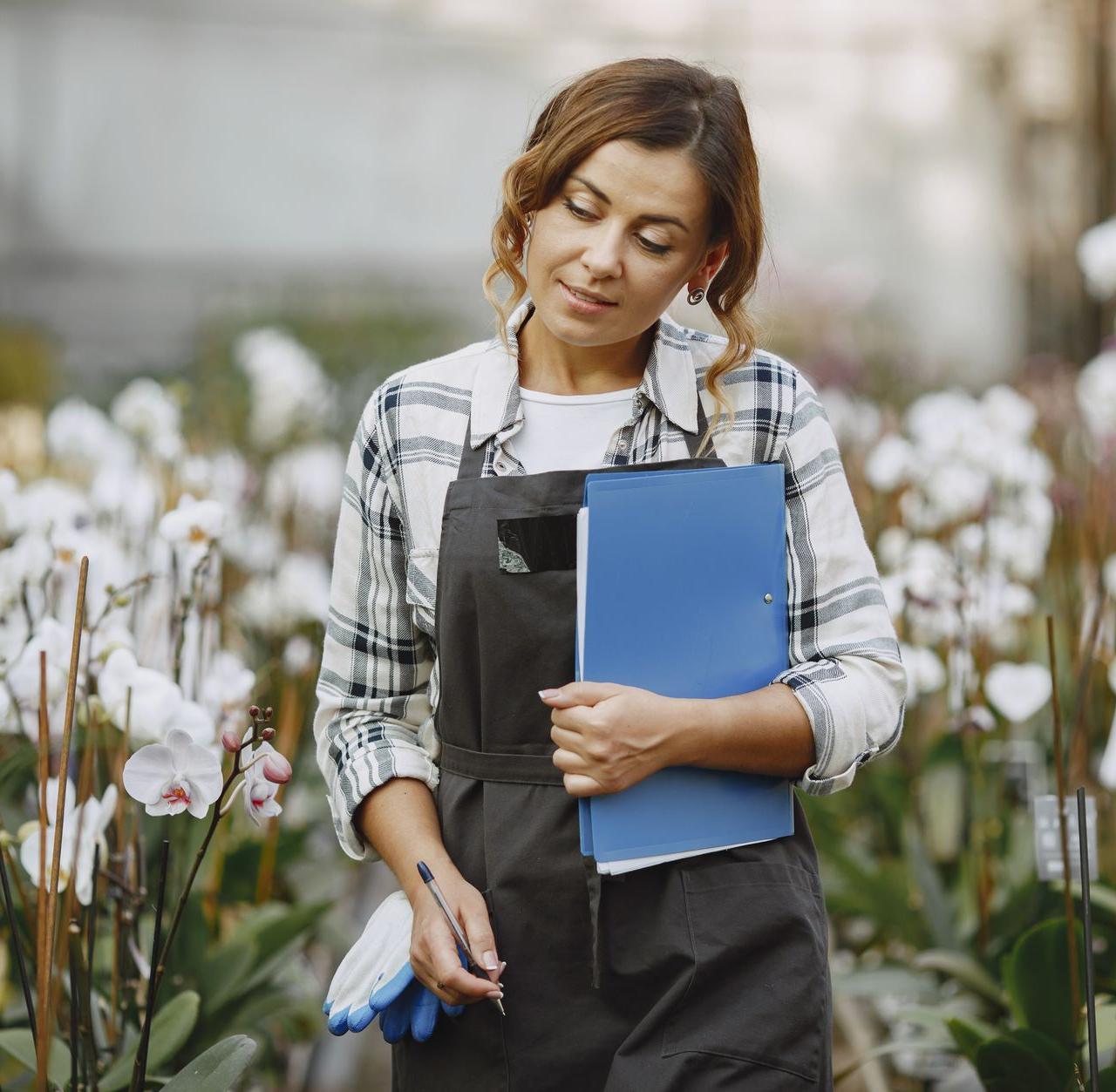Woman gardener in greenhouse holding a notebook, surrounded by blooming orchids.