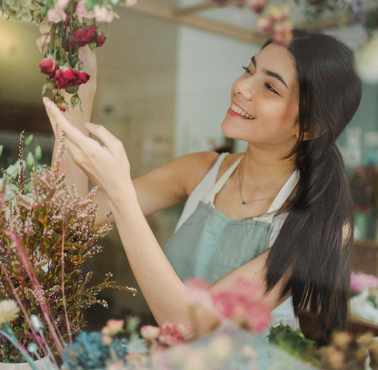 Young smiling dark haired female florist in apron arranging flowers while working in floral shop