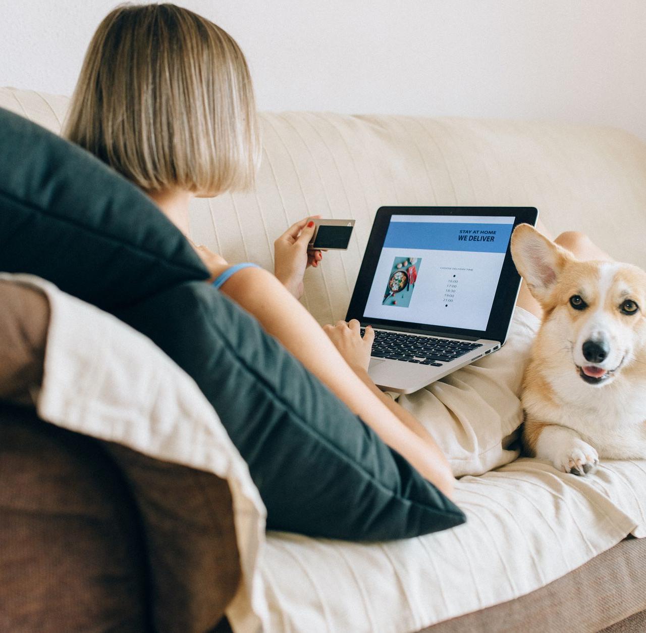 A woman using a laptop for online shopping with her corgi dog beside her on the couch.